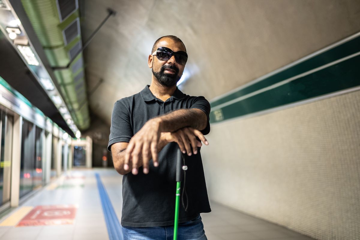 A blind man with medium skin, a shaved head and a beard poses casually while waiting for the subway. He is wearing sunglasses and rests his hands on top of his white cane.