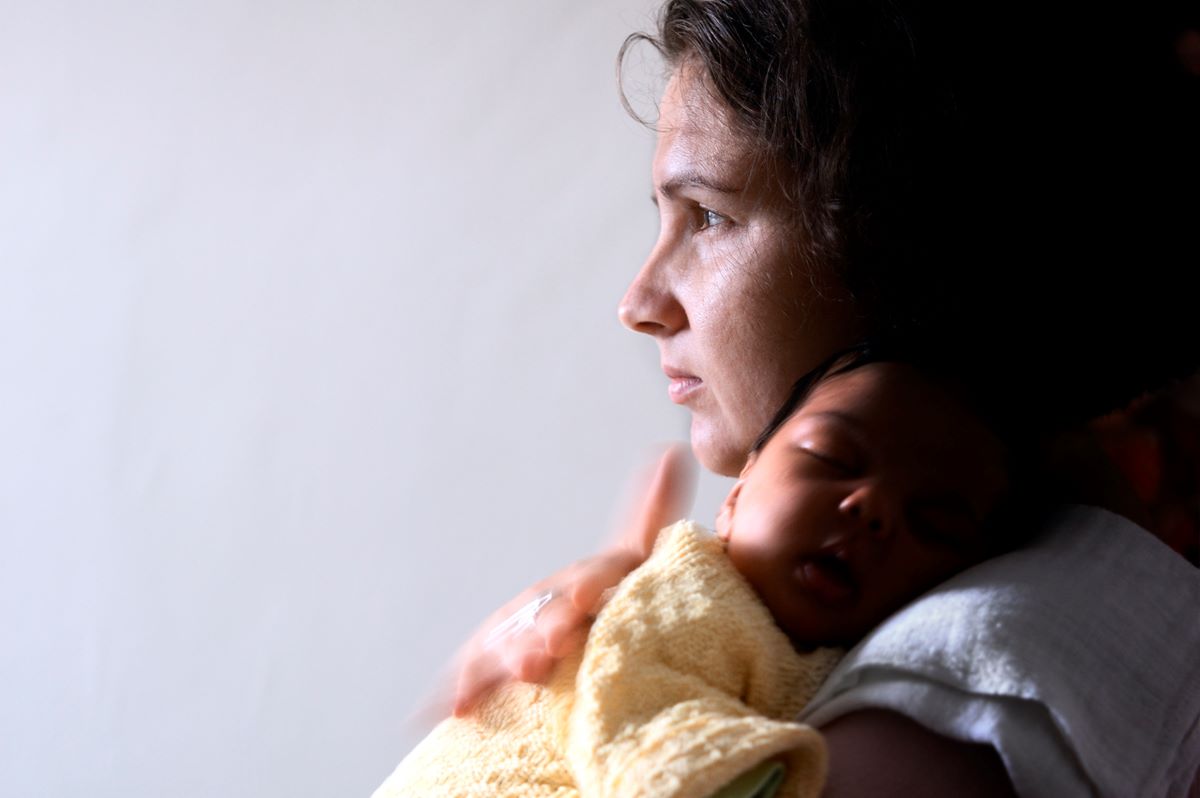 A worried mother looks out the window while holding her baby