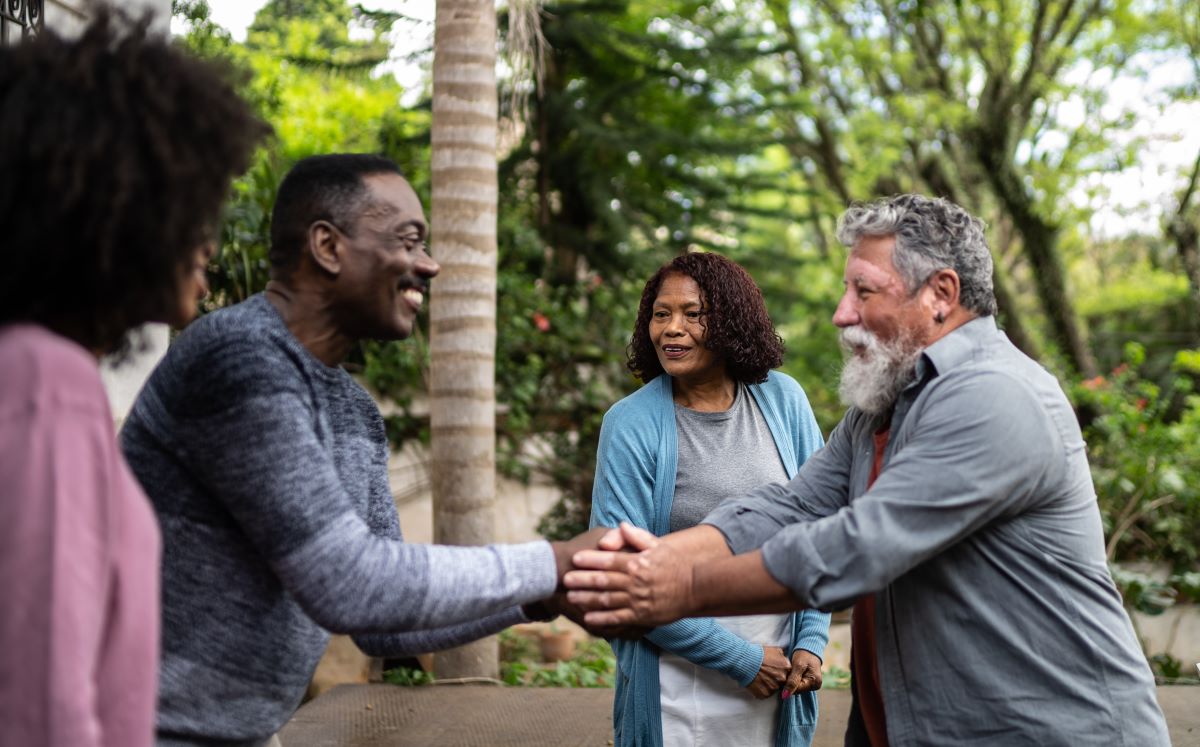Four people are meeting each other for the first time outdoors on a sunny day. A Black man and a Latino man shake hands in the center.