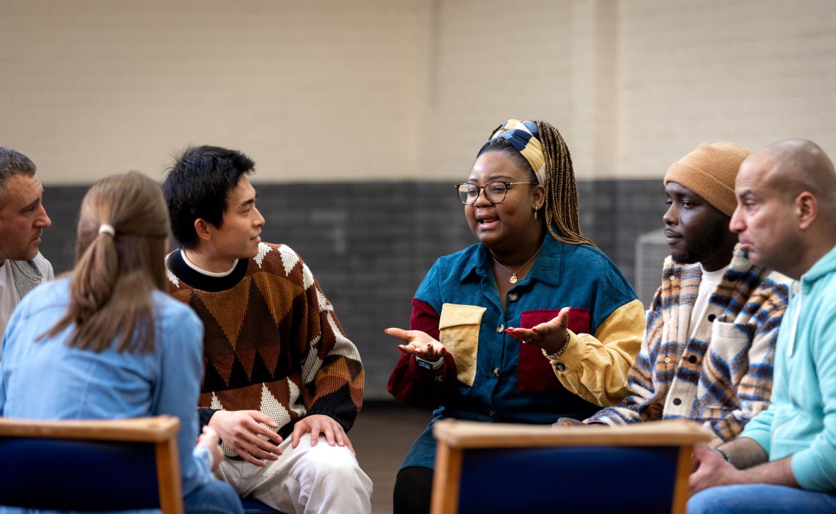 A diverse group of people sits in a circle discussing an issue. One person is talking and others are listening closely.