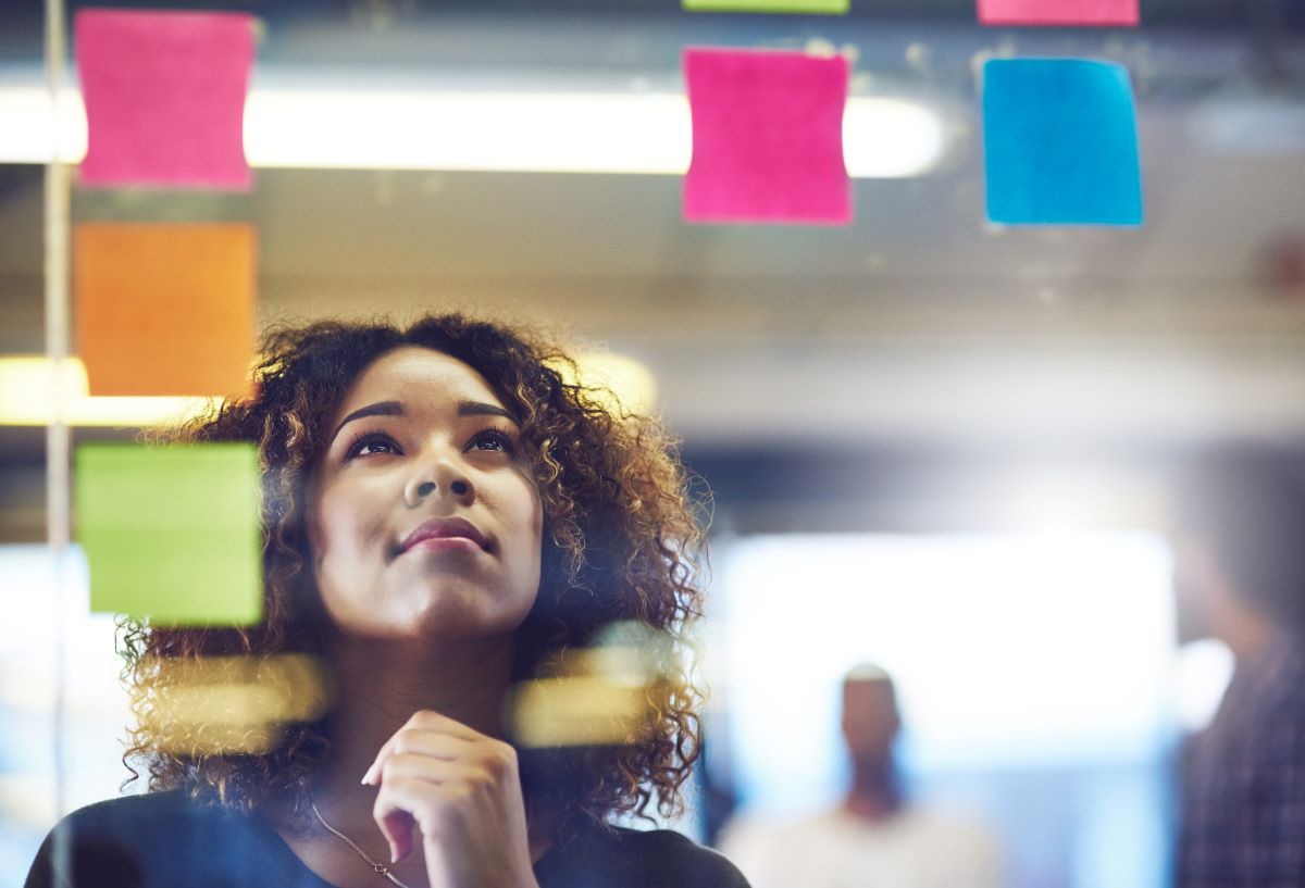 A Black woman looks up thoughtfully as she brainstorms using sticky notes on a glass wall with her colleagues in the background.