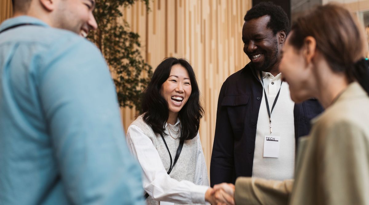 a smiling woman shakes hands with another woman while talking with colleagues at a networking event