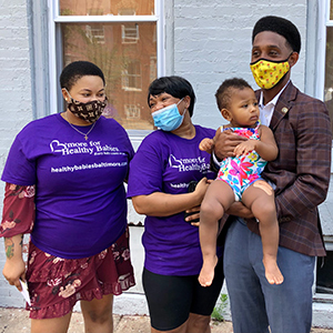 Image of people wearing B'more for Healthy Babies t-shirts and facemasks smiling and holding a toddler