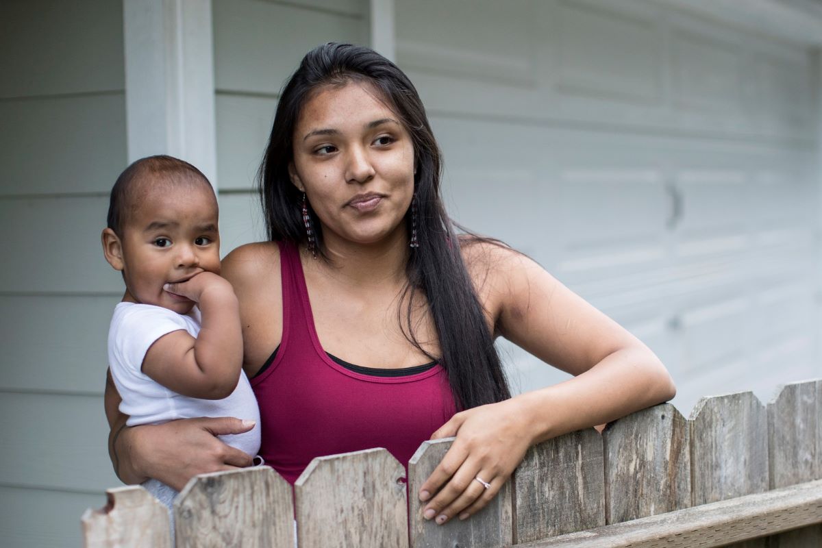 An Indigenous mother in warm weather clothing holds her infant on one hip while leaning over a fence