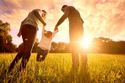 Believing to Becoming-58895064 Two parents hold their child's hands and lift her up in a field as the sun sets.