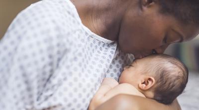 Maternal Health Equity A young African American mother in a hospital gown gently holds her infant in her arms and smiles down at her. The baby's eyes are closed.