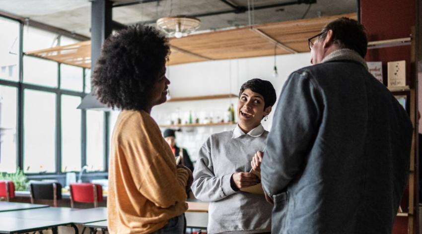 three people stand talking animatedly in a room with large windows