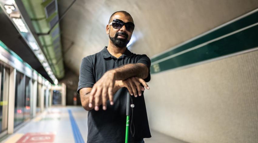 A blind man poses casually while waiting for the subway. He is wearing sunglasses and rests his hands on top of his green cane.