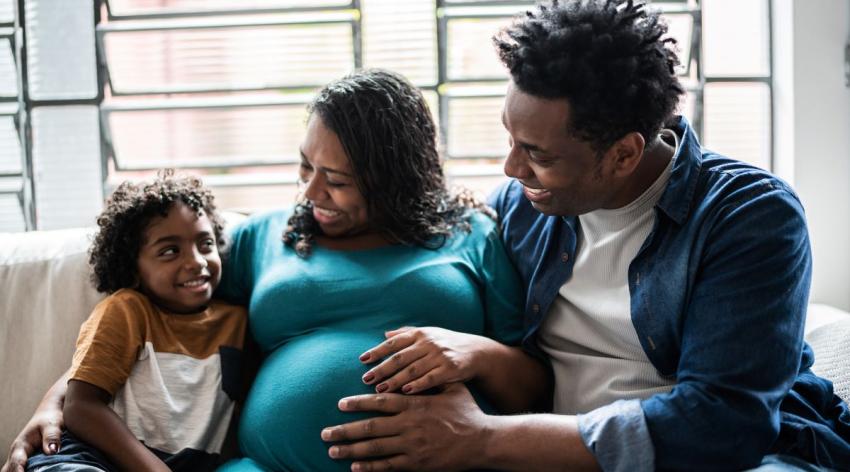 A Black mother and father excitedly tell their older child that he's going to have a sibling, while holding the mother's pregnant belly.