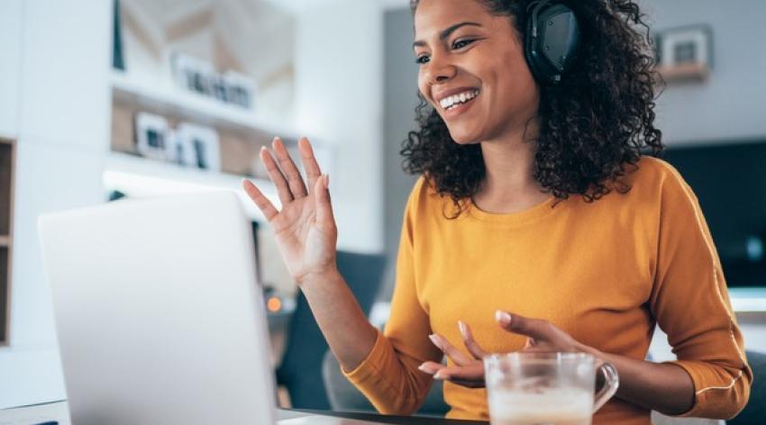 A Black woman with curly hair and headphones waves hello to colleagues via virtual meeting.