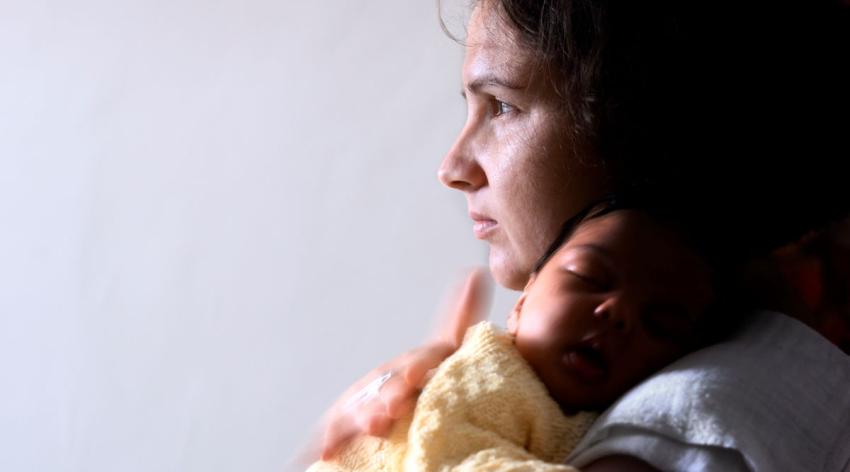 A worried mother looks out the window while holding her baby