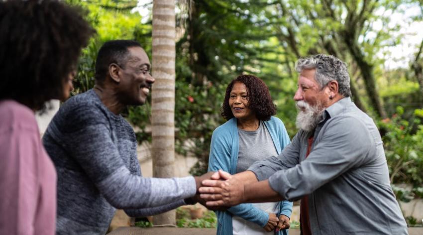 Four people are meeting each other for the first time outdoors on a sunny day. A Black man and a Latino man shake hands in the center.
