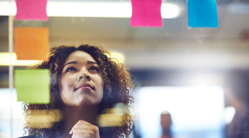 A Black woman looks up thoughtfully as she brainstorms using sticky notes on a glass wall with her colleagues in the background.