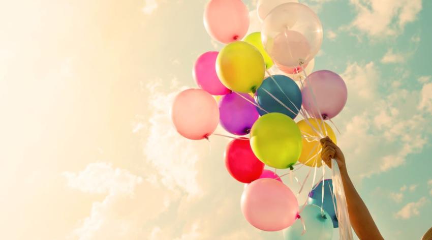 A hand holds a bouquet of colorful balloons against a background of a blue sky with fluffy clouds