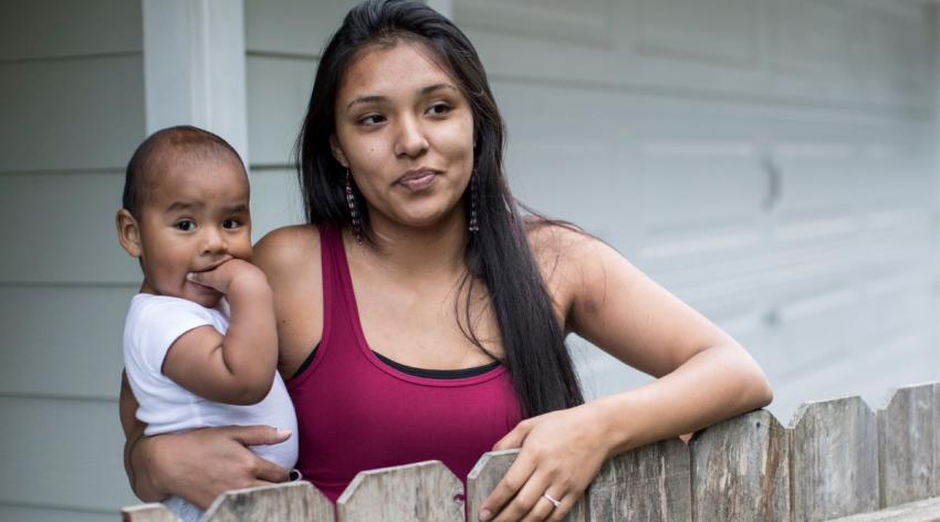 An Indigenous mother in warm weather clothing holds her infant while leaning over a fence